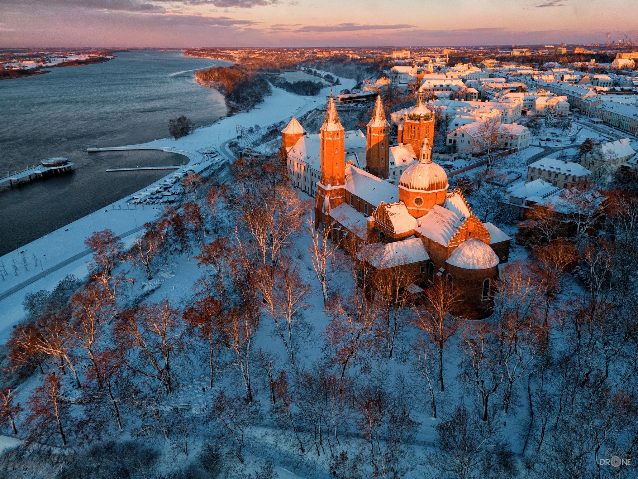 Zimowy krajobraz z lotu ptaka przedstawiający gotycką katedrę w Płocku oświetloną promieniami zachodzącego słońca, z widoczną rzeką Wisłą i ośnieżonym miastem w tle.
