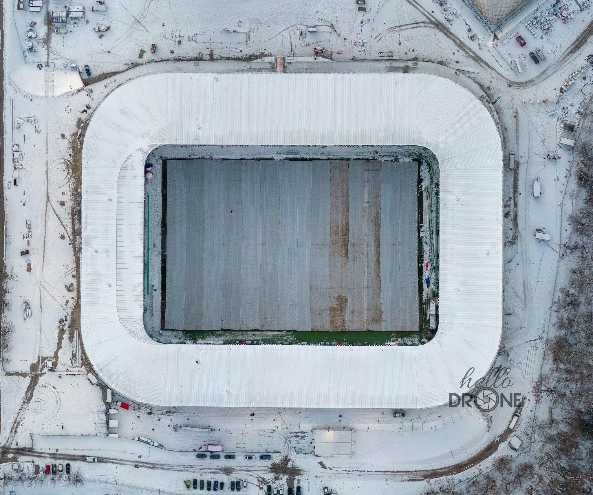 Widok z drona na zimowy stadion sportowy, pokryty śniegiem, z widocznymi śladami pojazdów na parkingu i wokół obiektu.