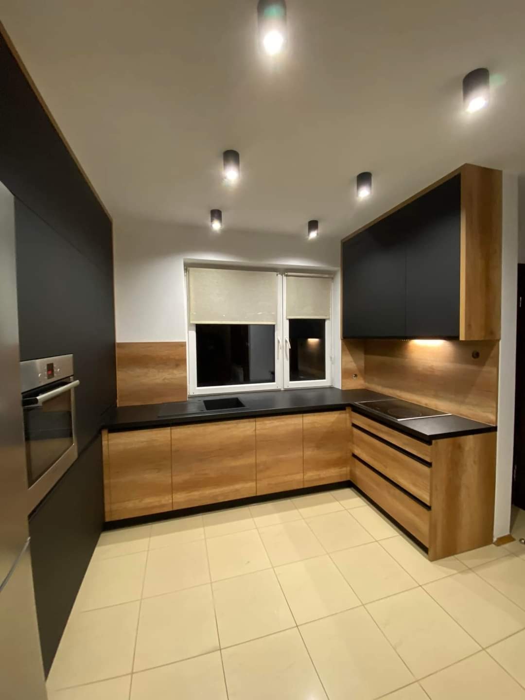 Nowoczesna kuchnia z drewnopodobnymi frontami szafek dolnych i czarnymi szafkami górnymi, czarny blat, oświetlenie punktowe na suficie i beżowa podłoga z płytek.