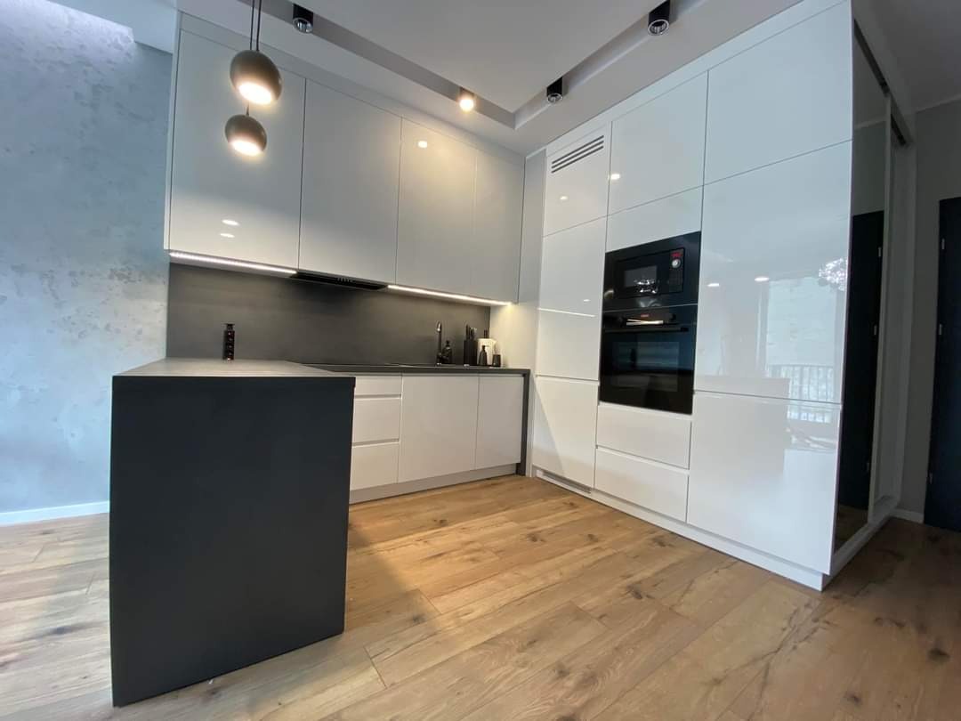 Nowoczesna kuchnia z białymi, błyszczącymi frontami szafek, czarnym blatem wyspy i drewnianą podłogą w odcieniach brązu, oświetlona wiszącymi lampami kulistymi.