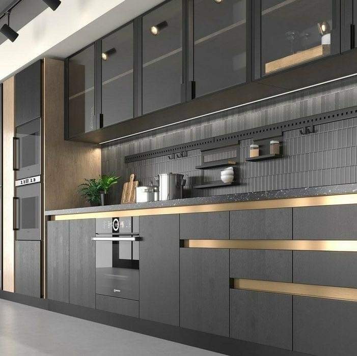 Nowoczesna kuchnia w odcieniach szarości z elementami złota, wyposażona w zabudowane sprzęty AGD i przeszklone szafki wiszące z oświetleniem punktowym.