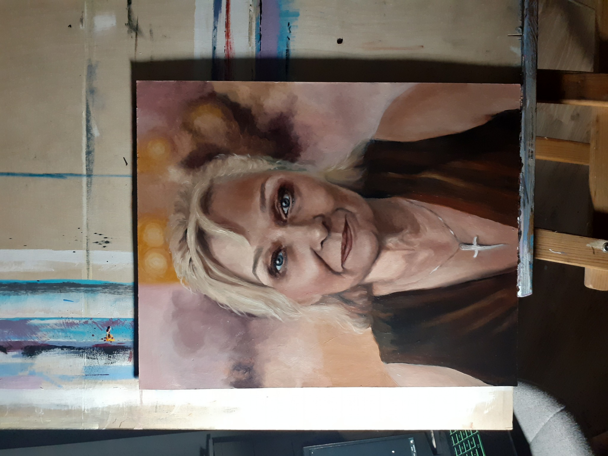 Portret olejny kobiety z blond włosami i srebrnym krzyżykiem na szyi, ustawiony na sztaludze w pracowni malarskiej.