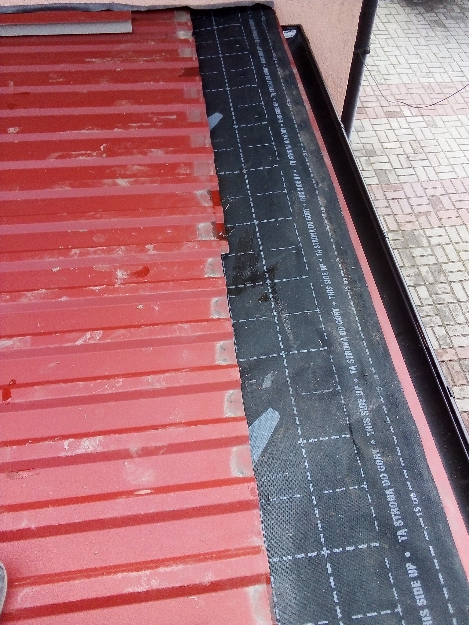 Czerwona blacha trapezowa na dachu z przyklejoną czarną membraną hydroizolacyjną z nadrukiem 'TA STRONA DO GÓRY' i podziałką co 15 cm, widoczne łączenia blachy i fragment rynny.