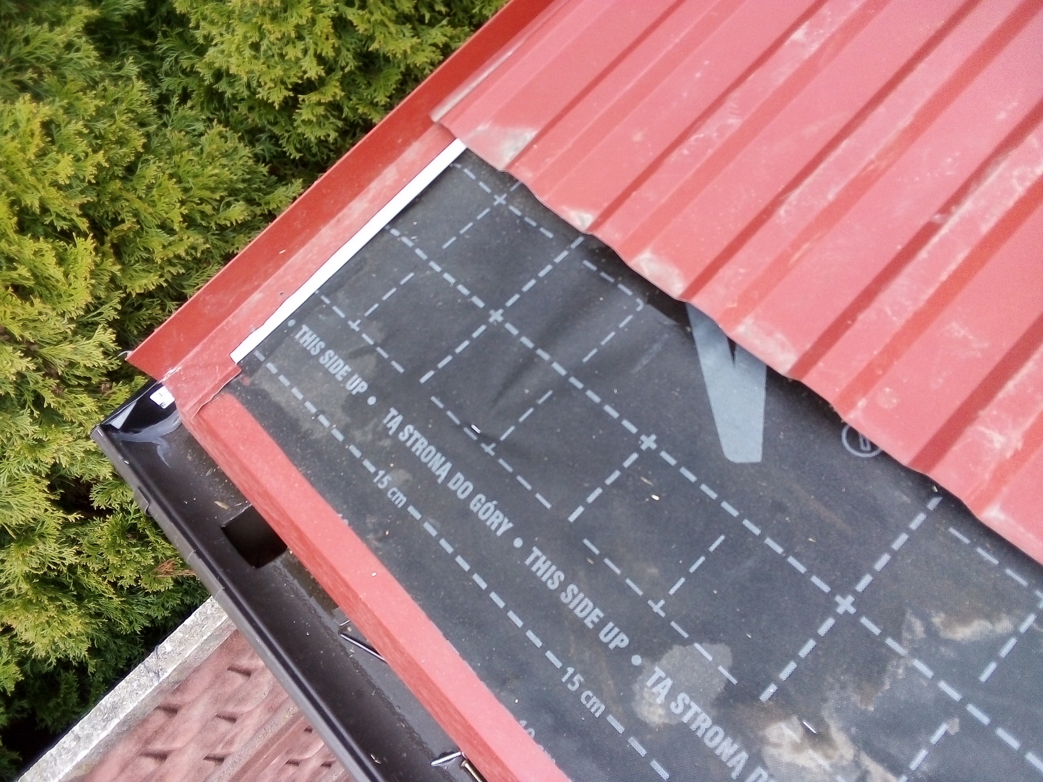 Fragment dachu z czerwoną blachodachówką i czarną membraną dachową z nadrukiem 'THIS SIDE UP / TA STRONA DO GORY', widoczny fragment rynny i zieleń w tle.
