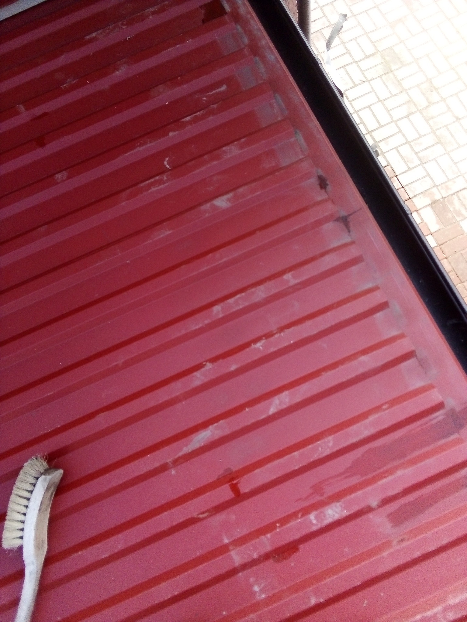 Czerwona, pomalowana powierzchnia z widocznymi smugami i zabrudzeniami, czyszczona szczotką z jasnym włosiem, fragment chodnika z kostki brukowej w tle.