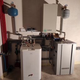 GoodTemp Klimatyzacja - Zestaw urządzeń grzewczych w kotłowni: biały zasobnik ciepłej wody COSMO, szara jednostka Auratsu z czynnikiem R32, naczynia przeponowe w kolorach niebieskim i szarym, połączone rurami.