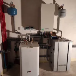 Zestaw urządzeń grzewczych w kotłowni: biały zasobnik ciepłej wody COSMO, szara jednostka Auratsu z czynnikiem R32, naczynia przeponowe w kolorach niebieskim i szarym, połączone rurami.