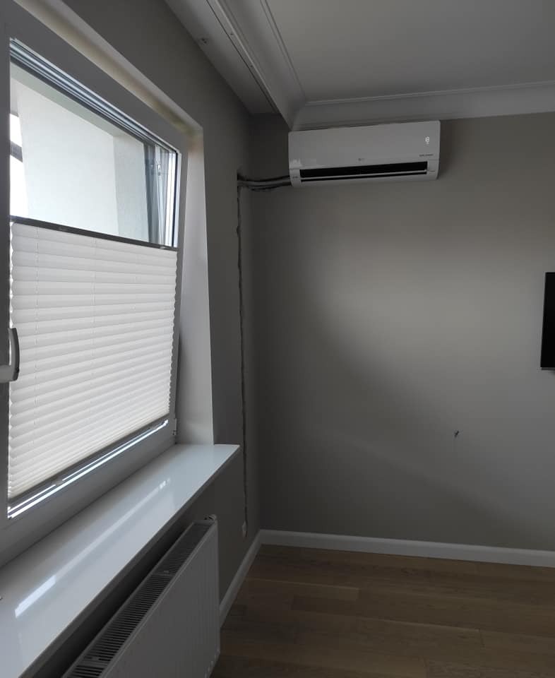 Biały klimatyzator ścienny zamontowany w szarym pokoju z drewnianą podłogą, widoczne przewody, roleta okienna plisowana.