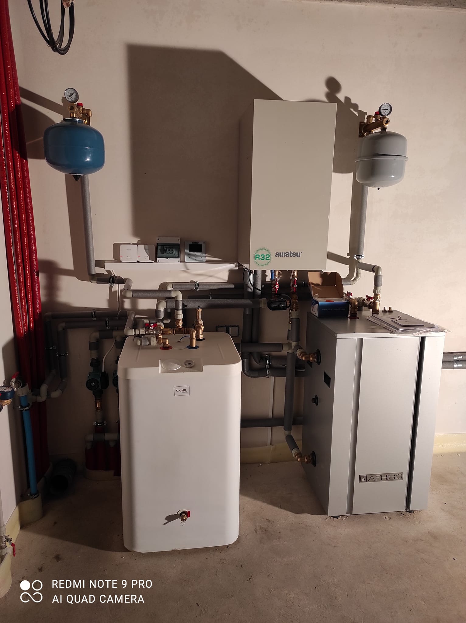 Zestaw urządzeń grzewczych w kotłowni: biały zasobnik ciepłej wody COSMO, szara jednostka Auratsu z czynnikiem R32, naczynia przeponowe w kolorach niebieskim i szarym, połączone rurami.