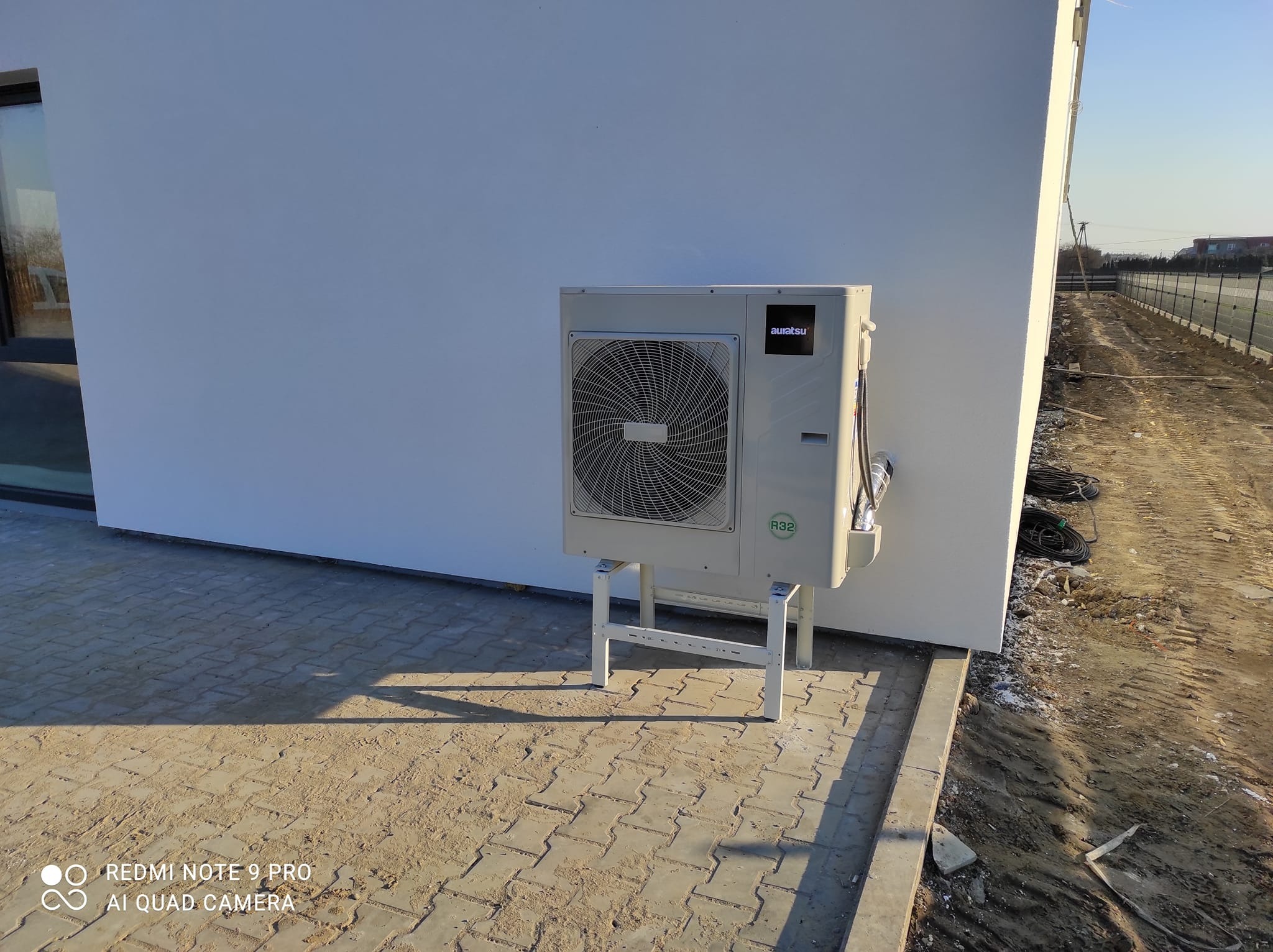 Zewnętrzna jednostka pompy ciepła marki Auratsu zamontowana na metalowym stelażu przy białej ścianie budynku, widoczny przewód i oznaczenie R32.