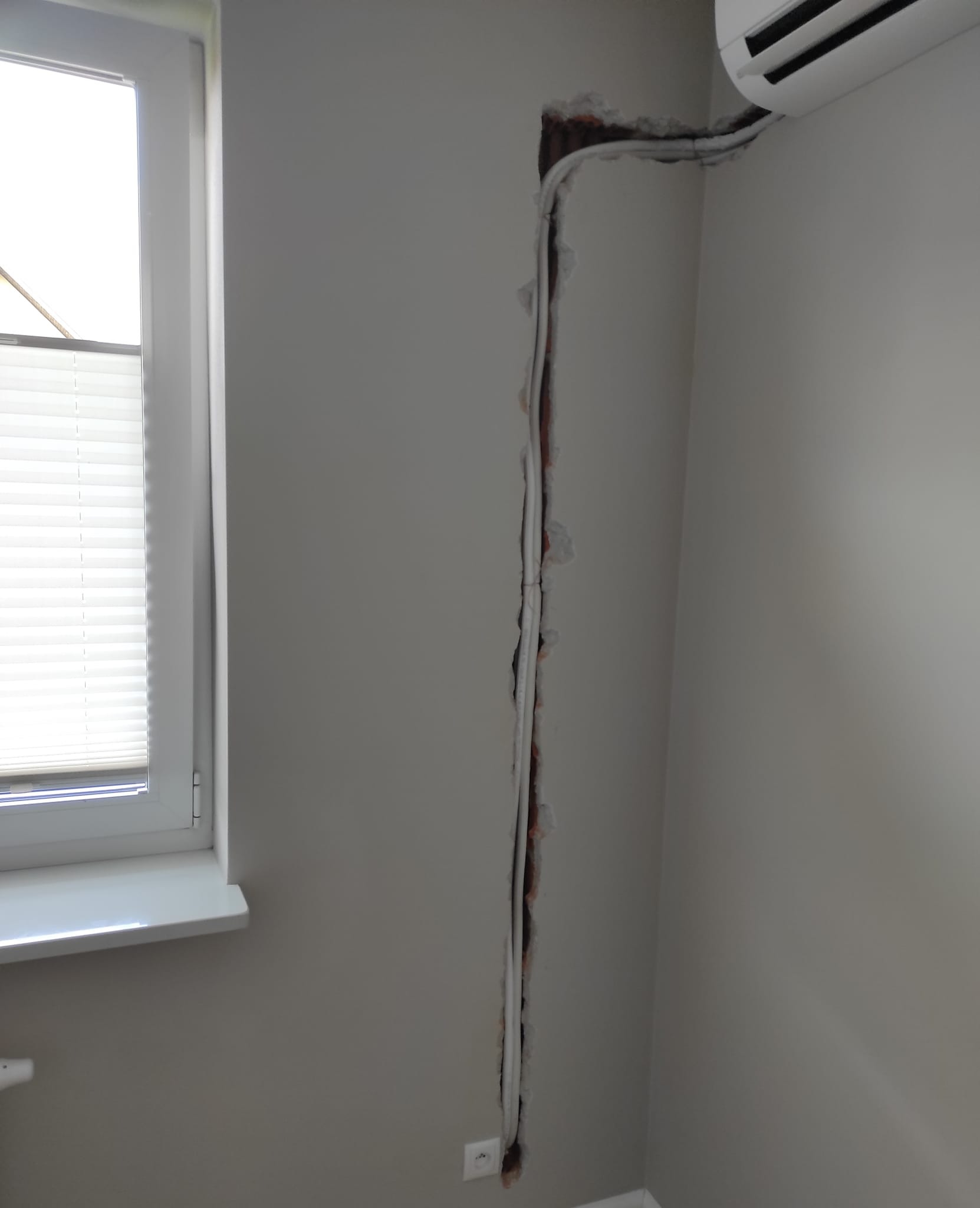 Montaż klimatyzacji w domu: frezowanie bruzdy w ścianie pod przewody, widoczny fragment klimatyzatora, okno z żaluzjami w tle.