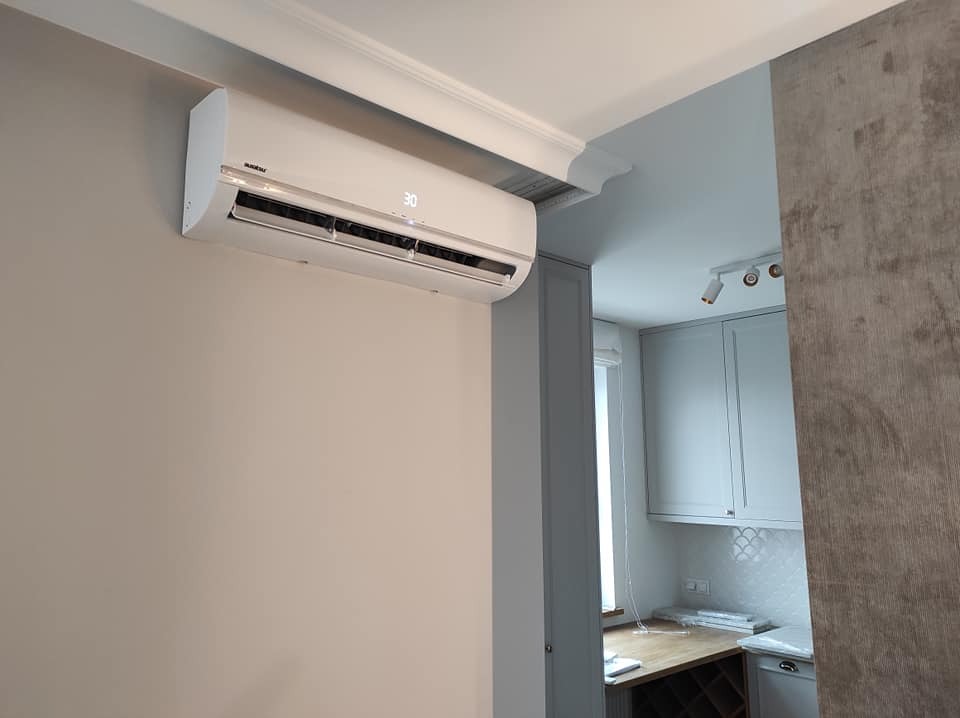 Biały klimatyzator zamontowany na ścianie w jasnym wnętrzu, widoczny wyświetlacz z temperaturą 30 stopni Celsjusza, dekoracyjny gzyms sufitowy.