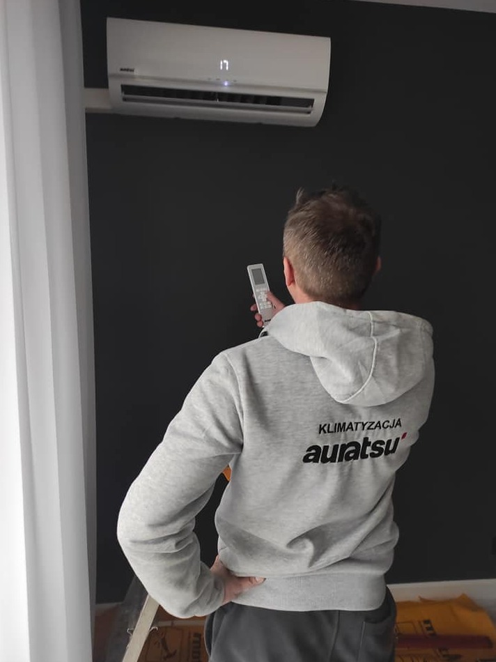 Mężczyzna w bluzie z napisem 'Klimatyzacja Auratsu' celuje pilotem w zamontowaną na ścianie klimatyzację. Na wyświetlaczu klimatyzatora widoczna temperatura 17 stopni. Ciemna ściana, jasna zasłona.
