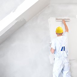 Projekt Home Adam Majchrowski - Mężczyzna w białym kombinezonie i żółtym kasku szpachluje ścianę w pokoju na poddaszu, używając dużej szpachelki; widoczne okno i surowe wykończenie.