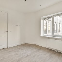 Projekt Home Adam Majchrowski - Puste, białe pomieszczenie po remoncie z panelami podłogowymi, grzejnikiem pod oknem i białymi drzwiami. Widoczne gniazdka elektryczne na ścianie.