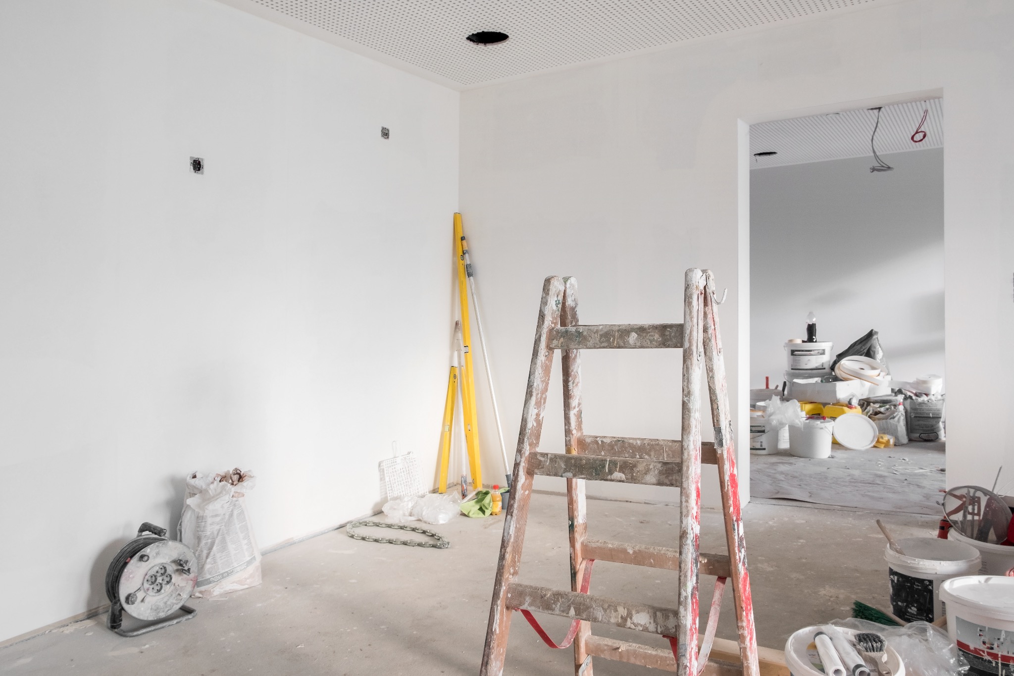 Wnętrze w trakcie prac wykończeniowych: białe ściany, perforowany sufit z otworem, drabina malarska z plamami farby na pierwszym planie, poziomice i torba z zaprawą w tle, widoczne puszki z farbą...