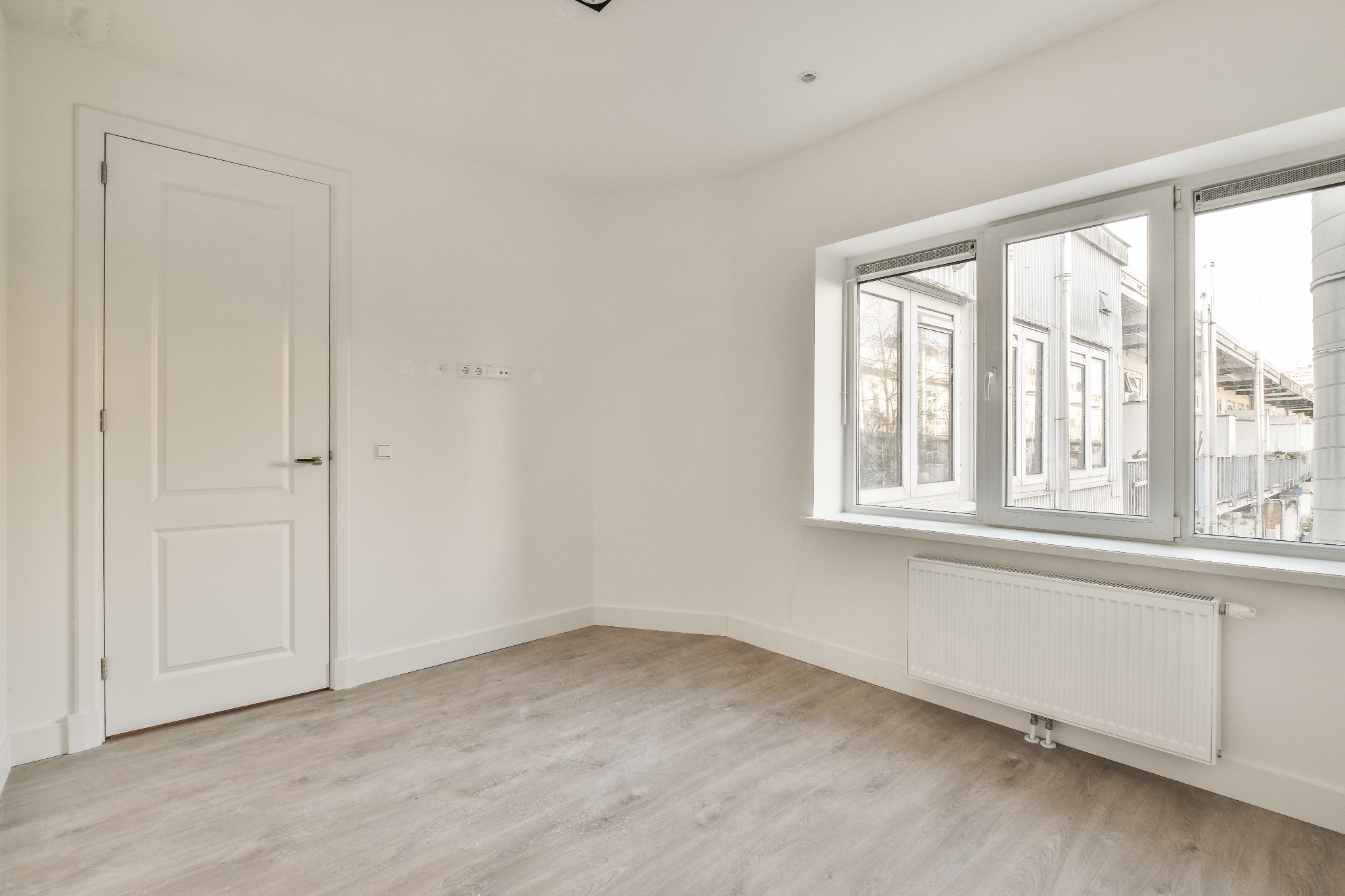 Puste, białe pomieszczenie po remoncie z panelami podłogowymi, grzejnikiem pod oknem i białymi drzwiami. Widoczne gniazdka elektryczne na ścianie.