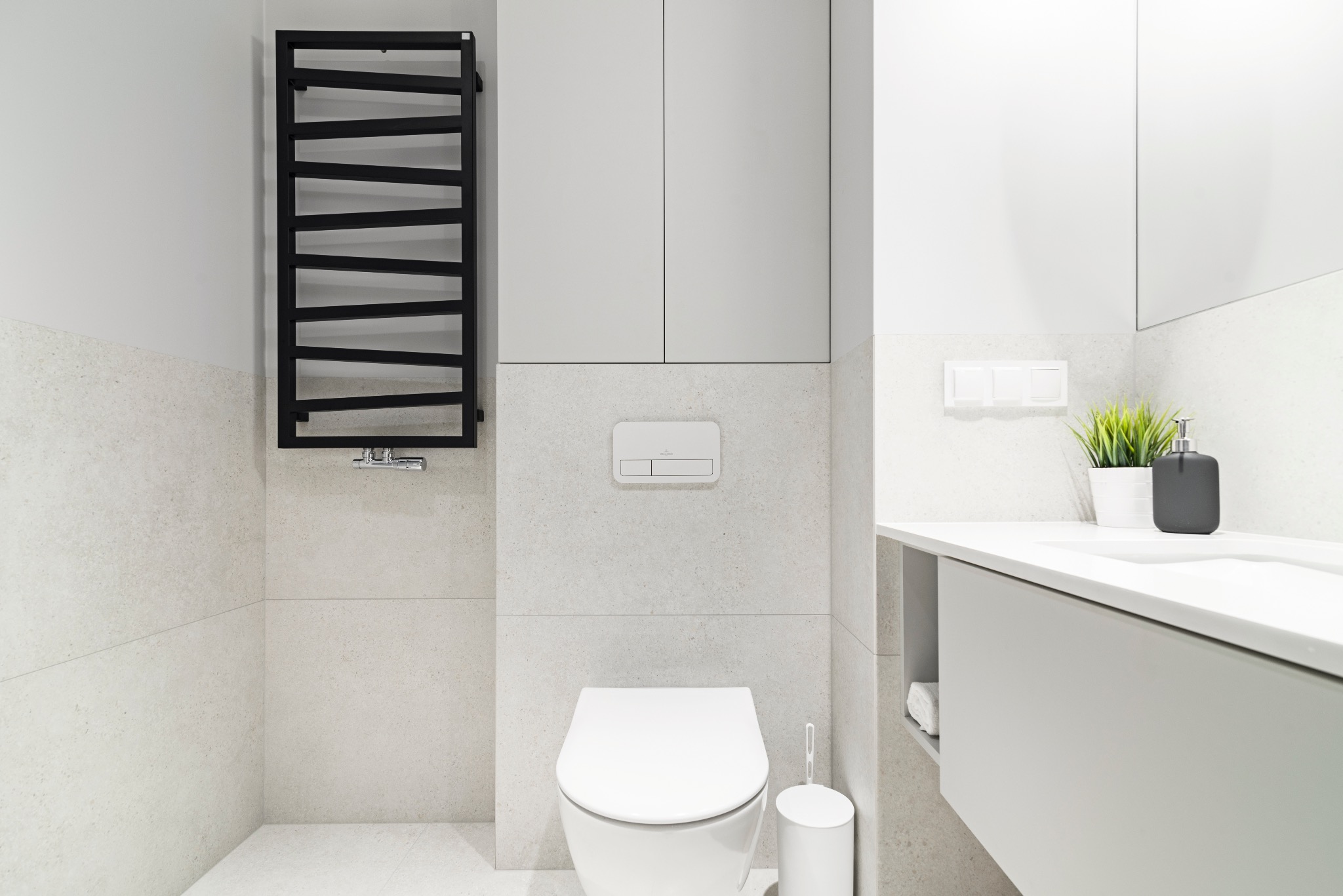 Minimalistyczna łazienka w odcieniach bieli i szarości z czarnym, drabinkowym grzejnikiem, wiszącą miską WC i umywalką z szafką, akcentem jest zielona roślina w doniczce.