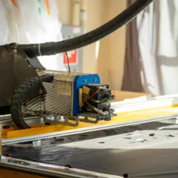 Automatyczna maszyna do cięcia materiałów z systemem odsysania, widoczna na stole roboczym z czarnym materiałem, w tle białe zasłony.