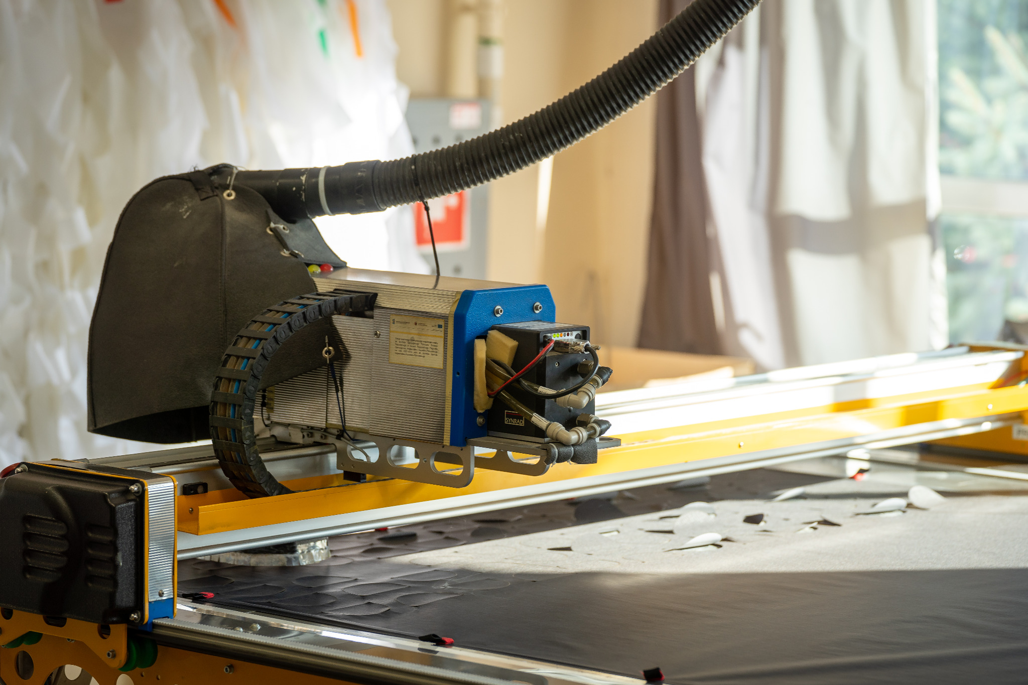 Automatyczna maszyna do cięcia materiałów z systemem odsysania, widoczna na stole roboczym z czarnym materiałem, w tle białe zasłony.