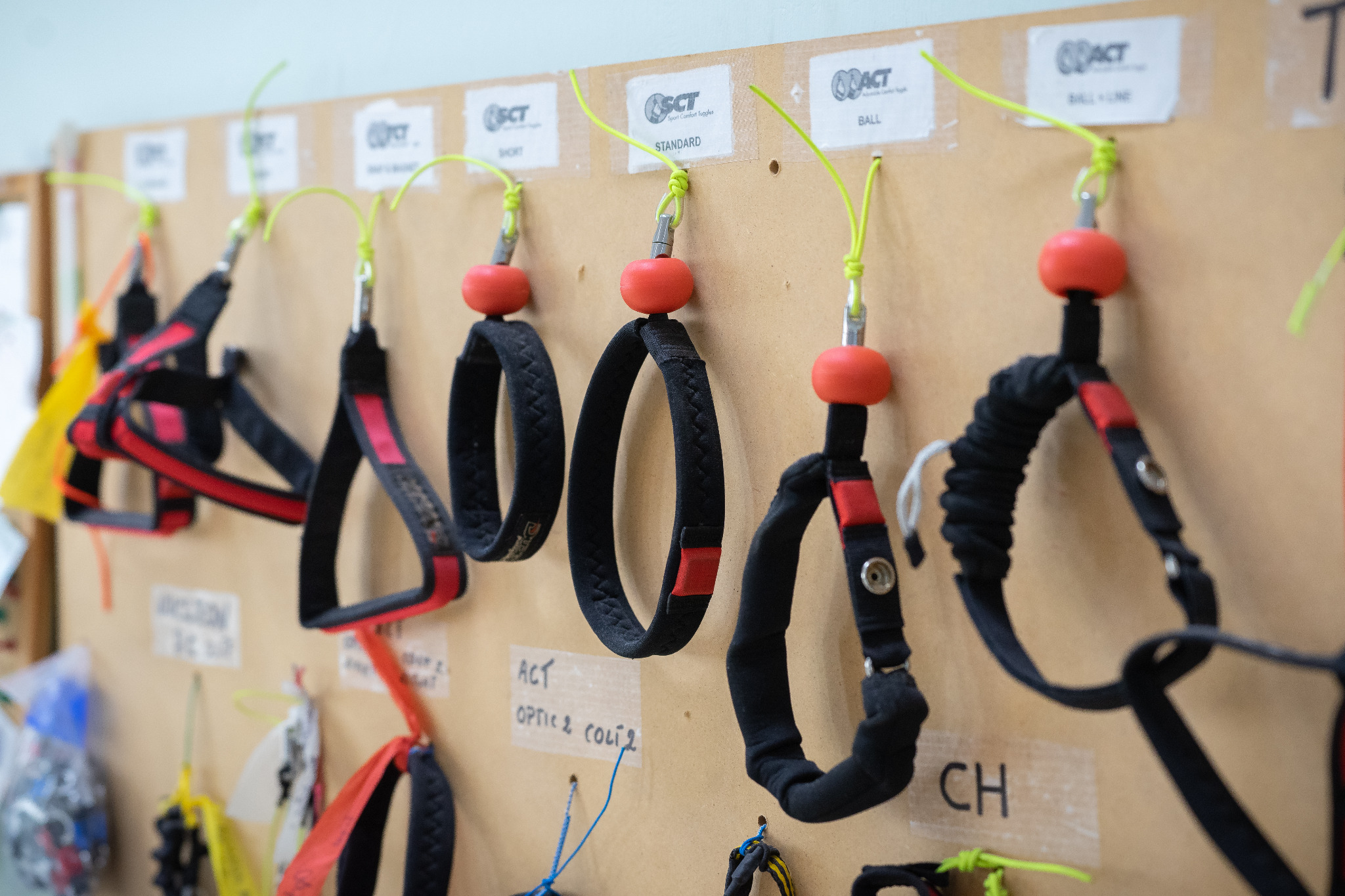 Ekspozycja różnych modeli pasków do okularów sportowych z czerwonymi elementami, zawieszonych na tablicy korkowej z etykietami informacyjnymi.