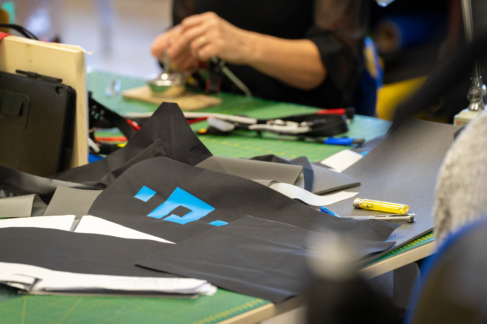 Fragment warsztatu z rozłożonymi kawałkami czarnego materiału z niebieskim nadrukiem, widoczne narzędzia, ręce pracownika w tle przy pracy.