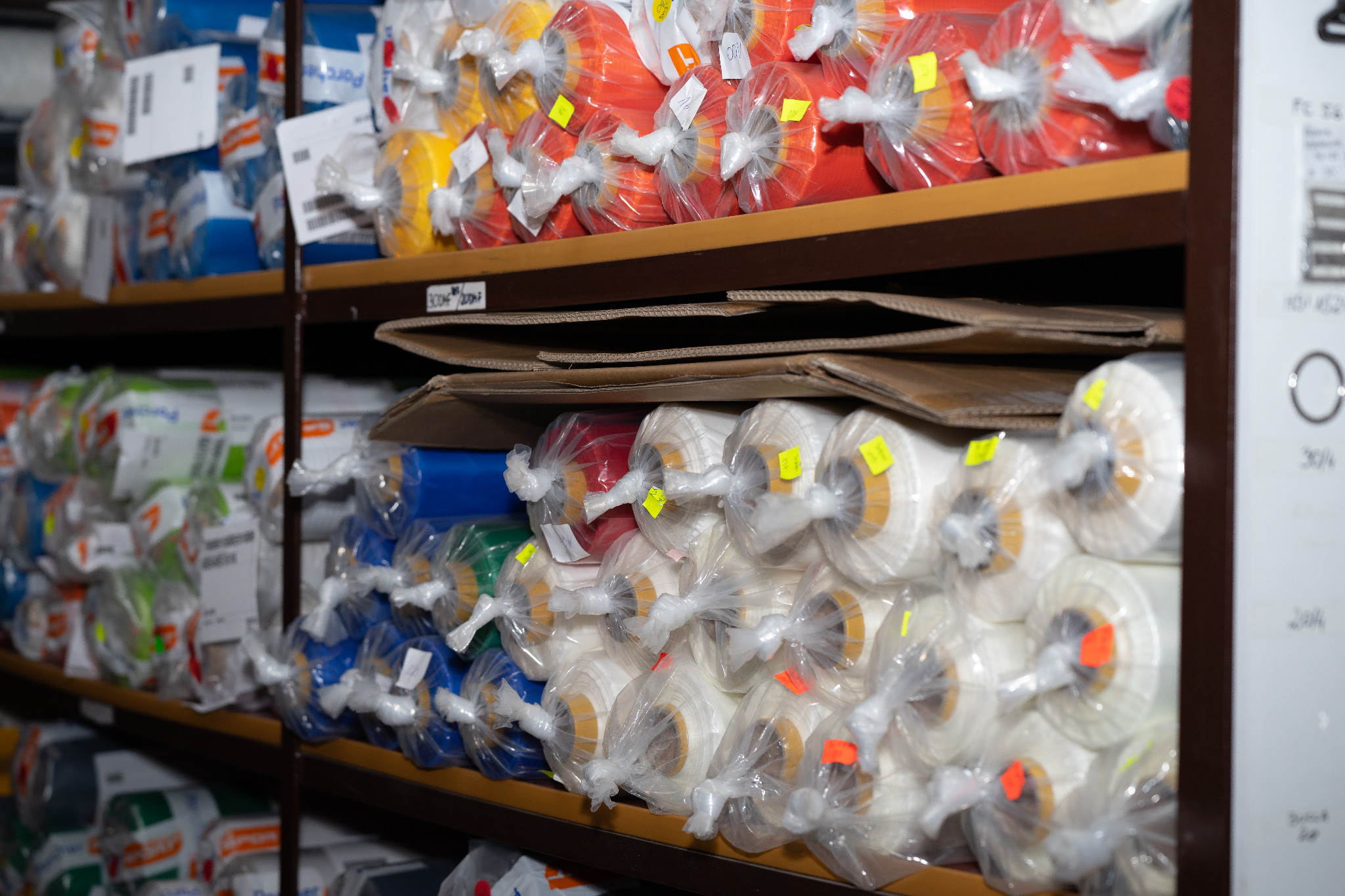 Regał magazynowy wypełniony kolorowymi rolkami tkanin zabezpieczonych folią i etykietami, widok z perspektywy dolnej półki.