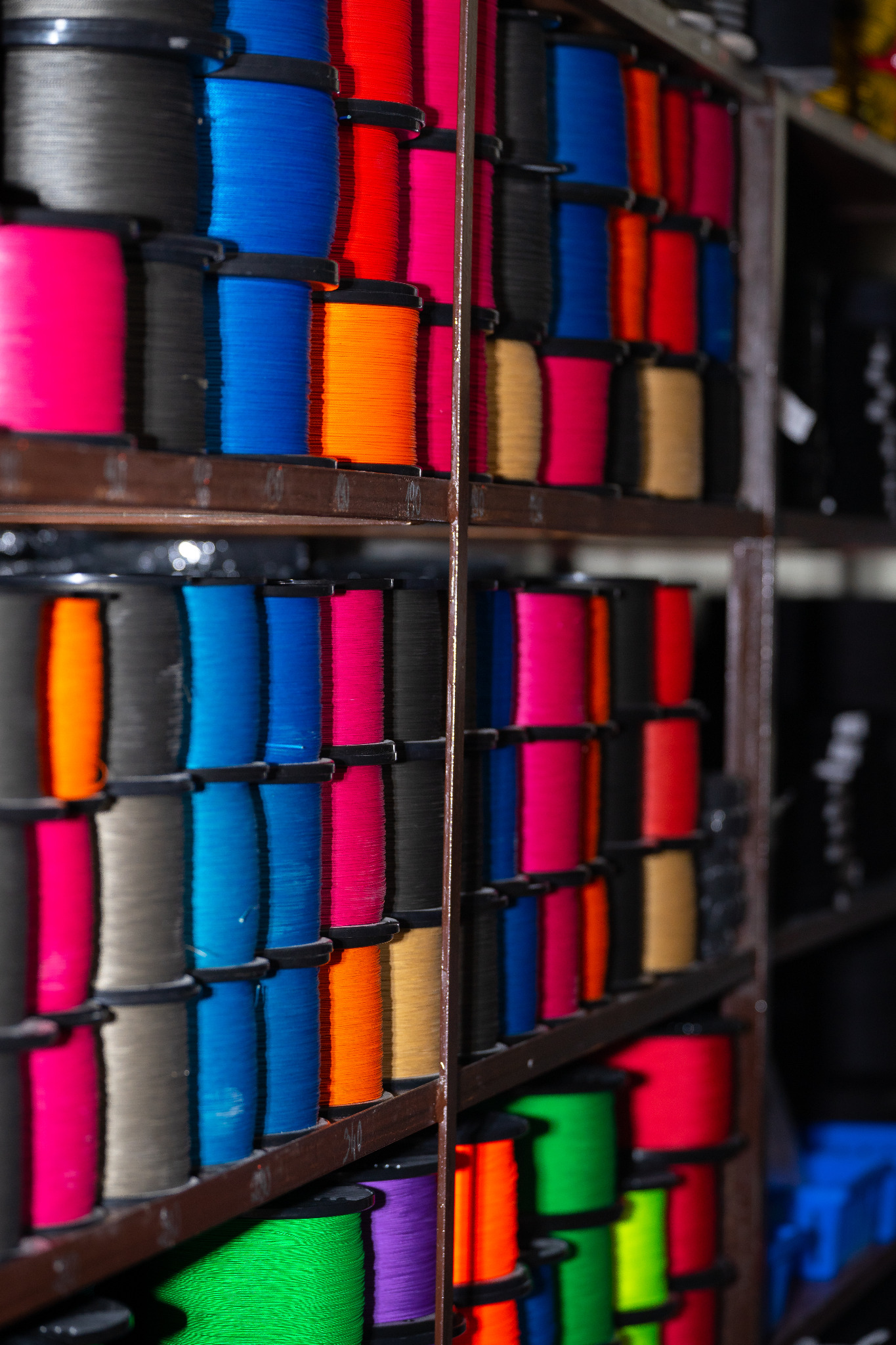 Regał z wieloma szpulami nici w różnych kolorach: różowe, niebieskie, pomarańczowe, czarne, czerwone, zielone i fioletowe, ułożone rzędami na ciemnych półkach.