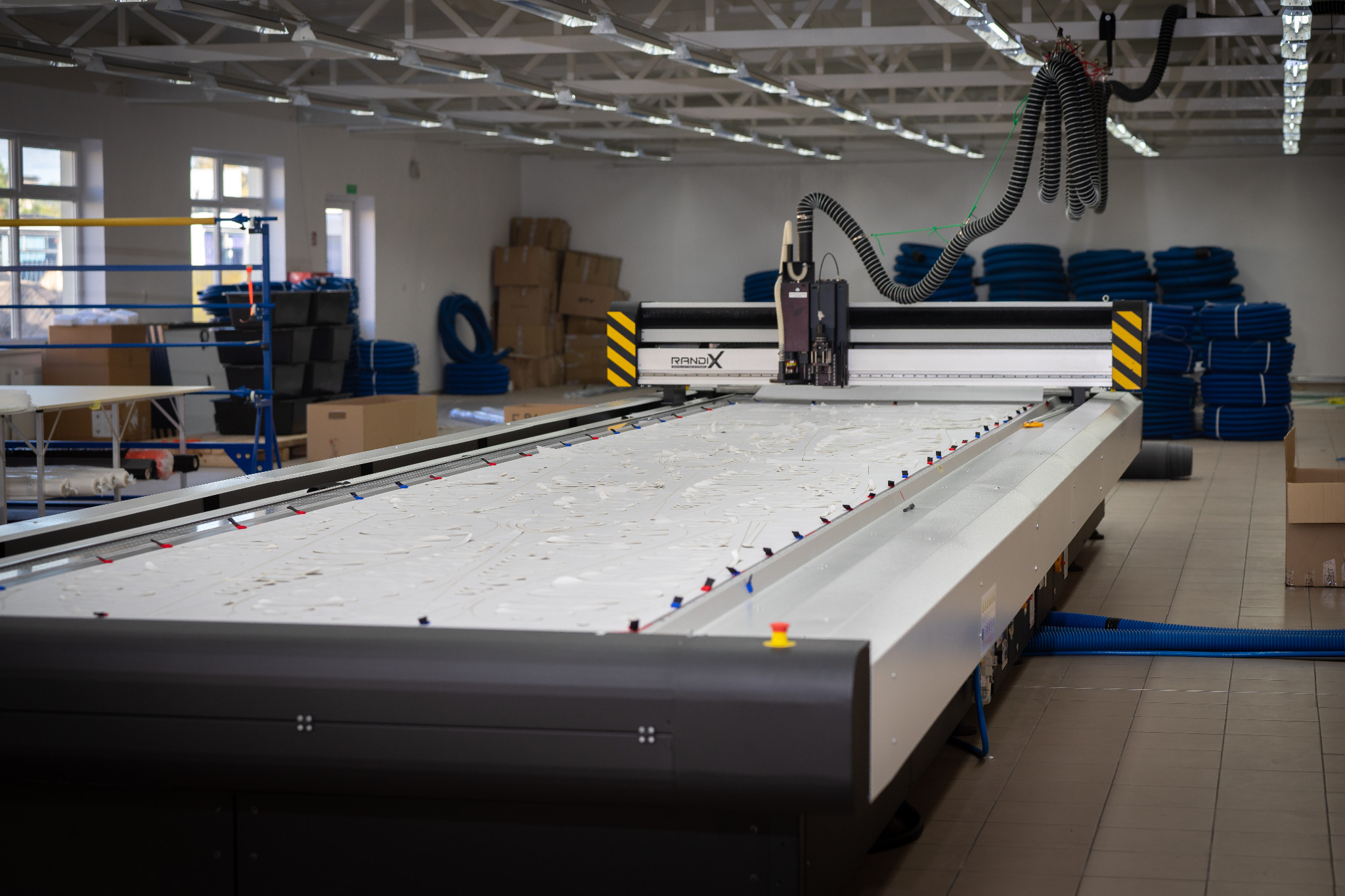 Duży ploter tnący z głowicą tnącą nad białą tkaniną, widoczne niebieskie rury i rolki materiału w tle, oświetlenie przemysłowe.