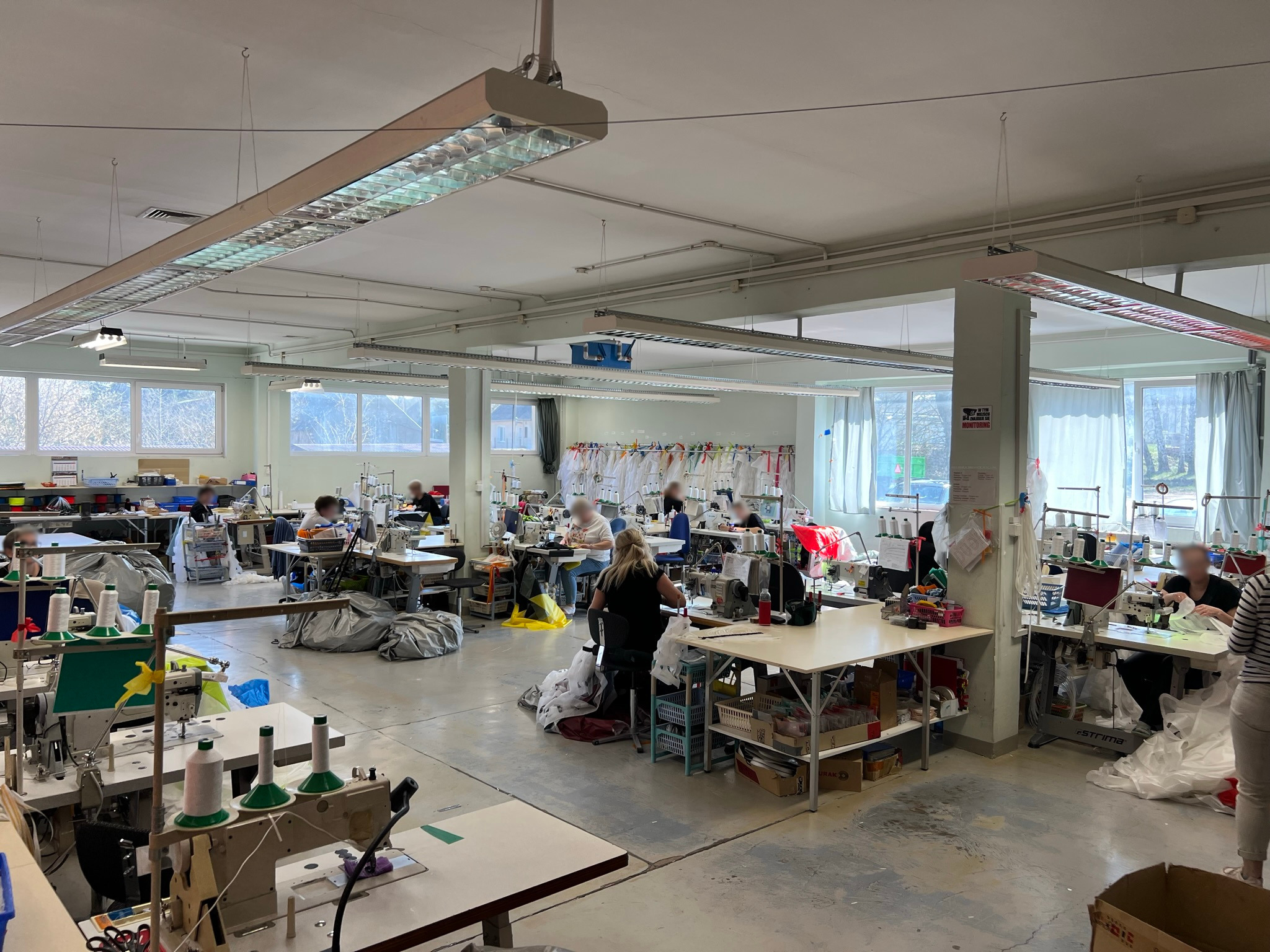 Przestronna hala produkcyjna z rzędami stanowisk do szycia, wyposażonych w maszyny i akcesoria, przy oknach wiszą gotowe wyroby tekstylne, a pracownicy w skupieniu wykonują swoje zadania.