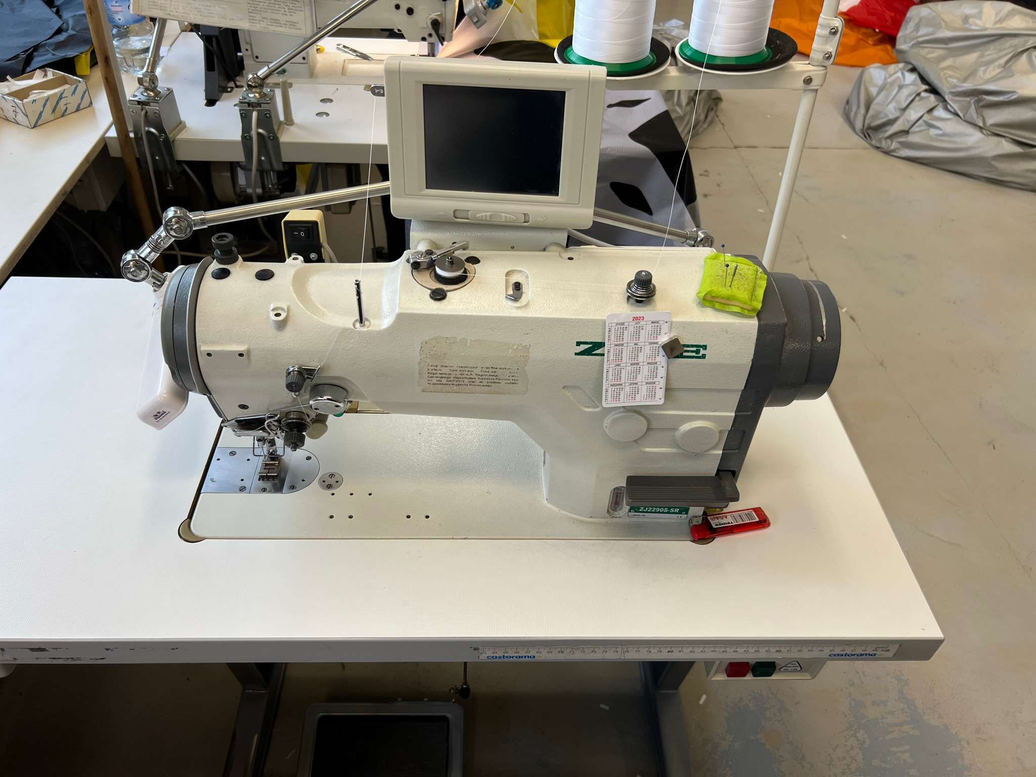 Profesjonalna maszyna szwalnicza ZUKI z wyświetlaczem, igłami i podkładką na igły, ustawiona na stole z miarką Castorama, w tle rolki nici i materiały.