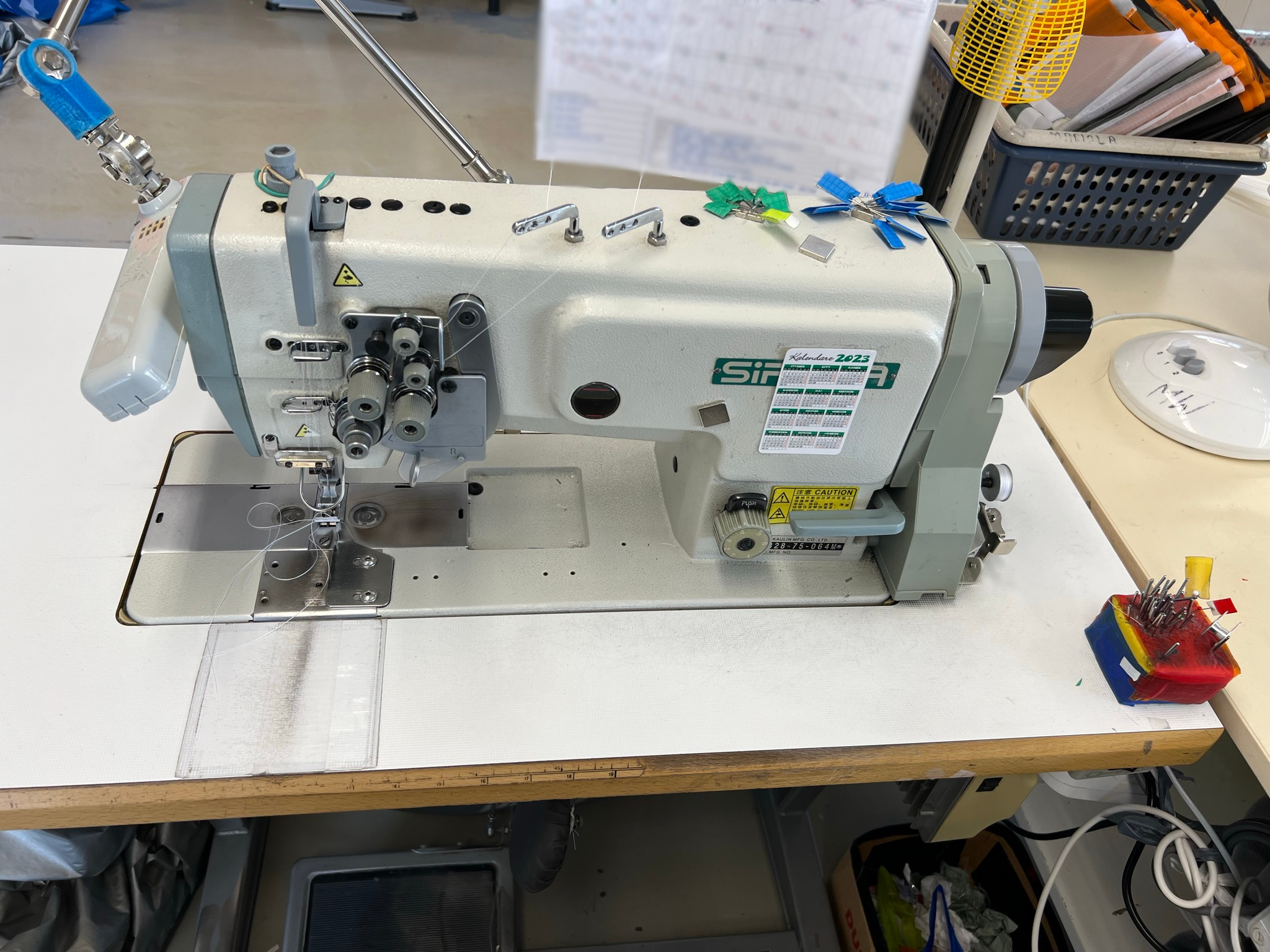 Profesjonalna maszyna do szycia przemysłowego z nawleczoną nicią, widoczne akcesoria krawieckie, takie jak szpilki i znaczniki, kalendarz firmowy na obudowie.