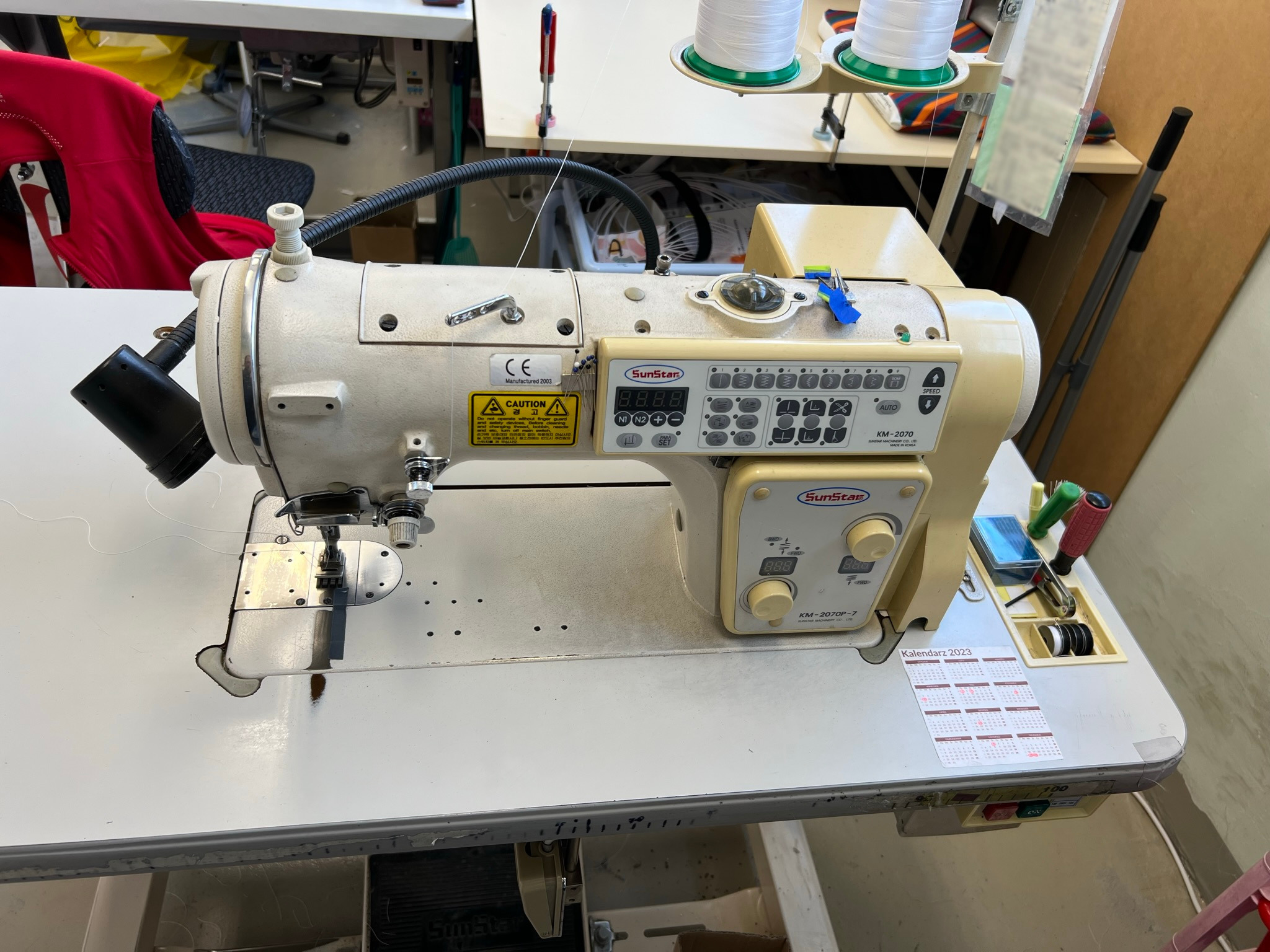 Profesjonalna maszyna szwalnicza SunStar KM-2070 na stole roboczym z kalendarzem 2023, widoczne szpule nici i narzędzia, oświetlenie warsztatowe.