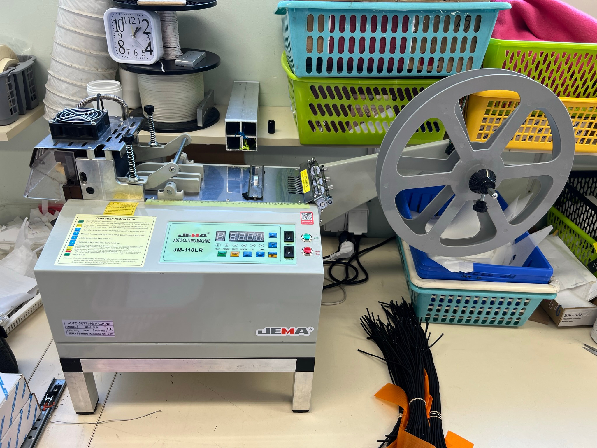Automatyczna maszyna do cięcia taśm JEMA JM-110LR w szwalni, z widocznym panelem sterowania, szpulą taśmy i gotowymi pęczkami czarnych taśm z pomarańczowymi akcentami.