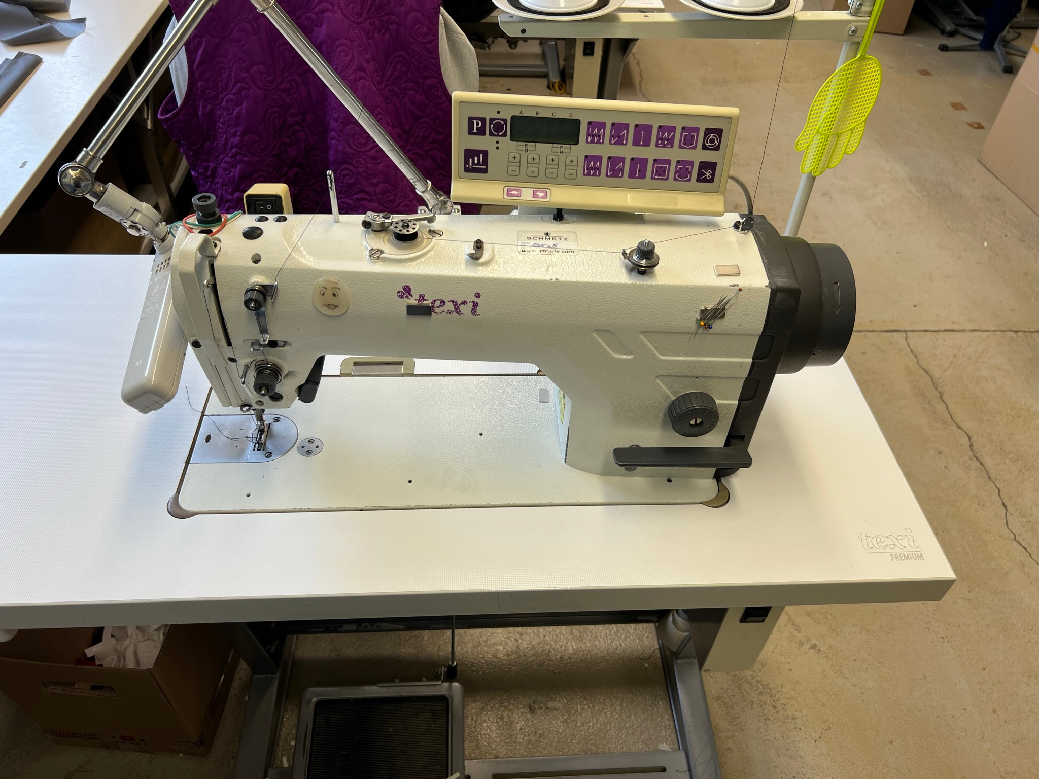 Profesjonalna, biała maszyna do szycia przemysłowego marki Texi Premium z panelem sterowania i lampką, ustawiona na białym stole w szwalni. Na maszynie naklejki dekoracyjne.
