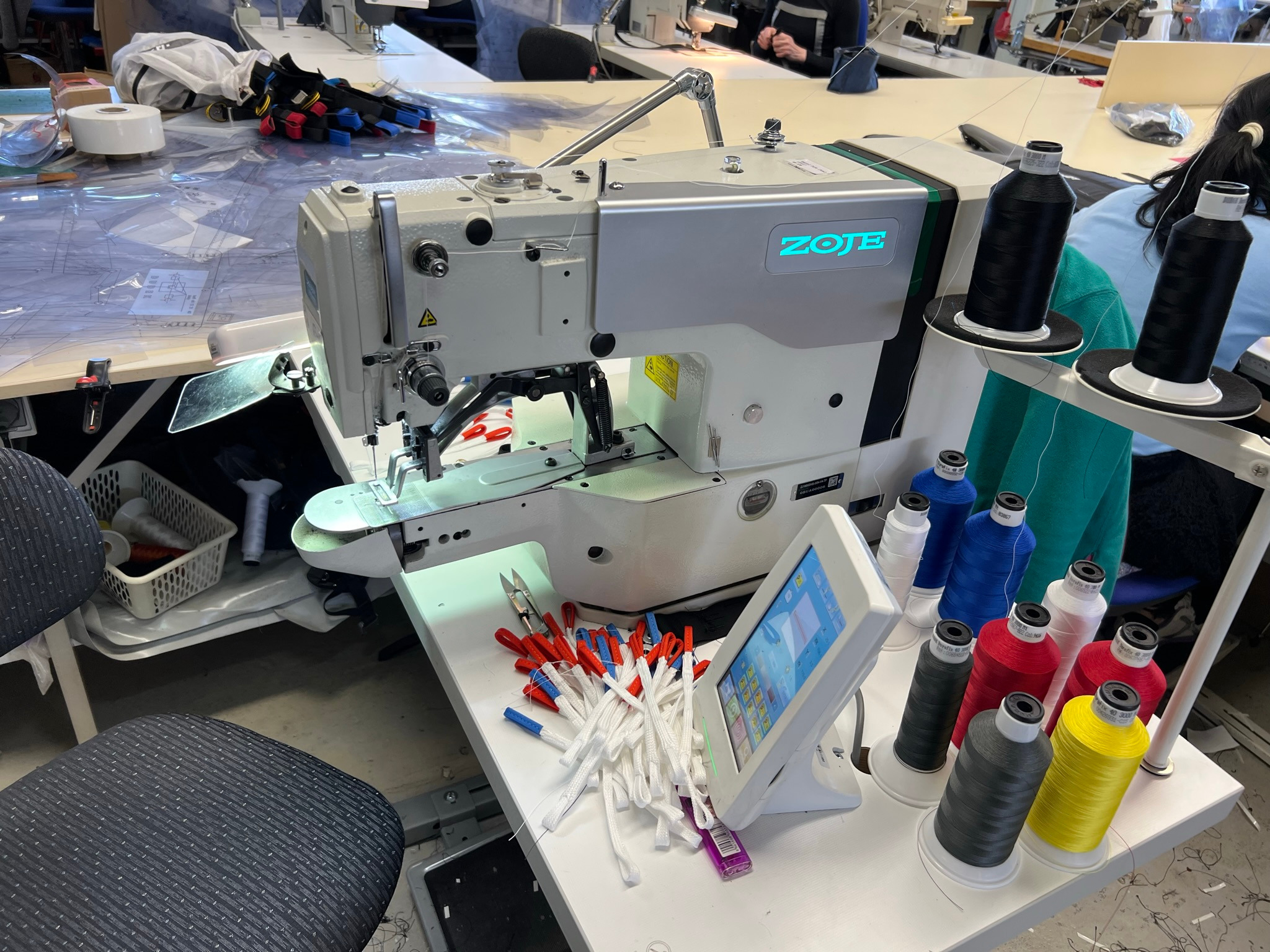 Przemysłowa maszyna do szycia Zoję z ekranem dotykowym, obok leżą pęczki sznurków z końcówkami w kolorach flagi Polski, nici w różnych kolorach stoją w rzędzie, w tle widoczny stół z wykrojami.