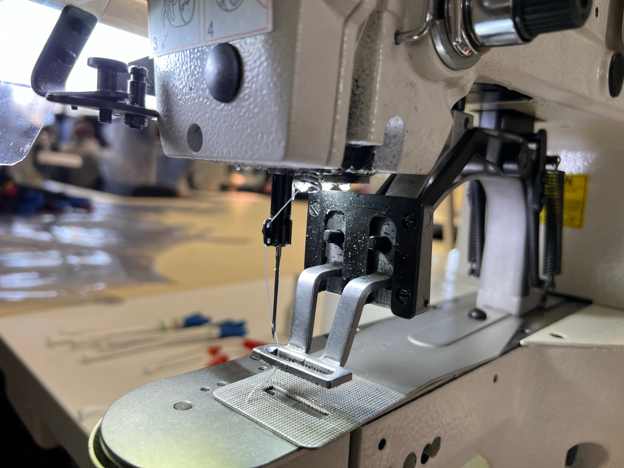 Zbliżenie na stopkę i igłę przemysłowej maszyny do szycia, widoczne detale mechanizmu oraz nitka, w tle stół z akcesoriami.