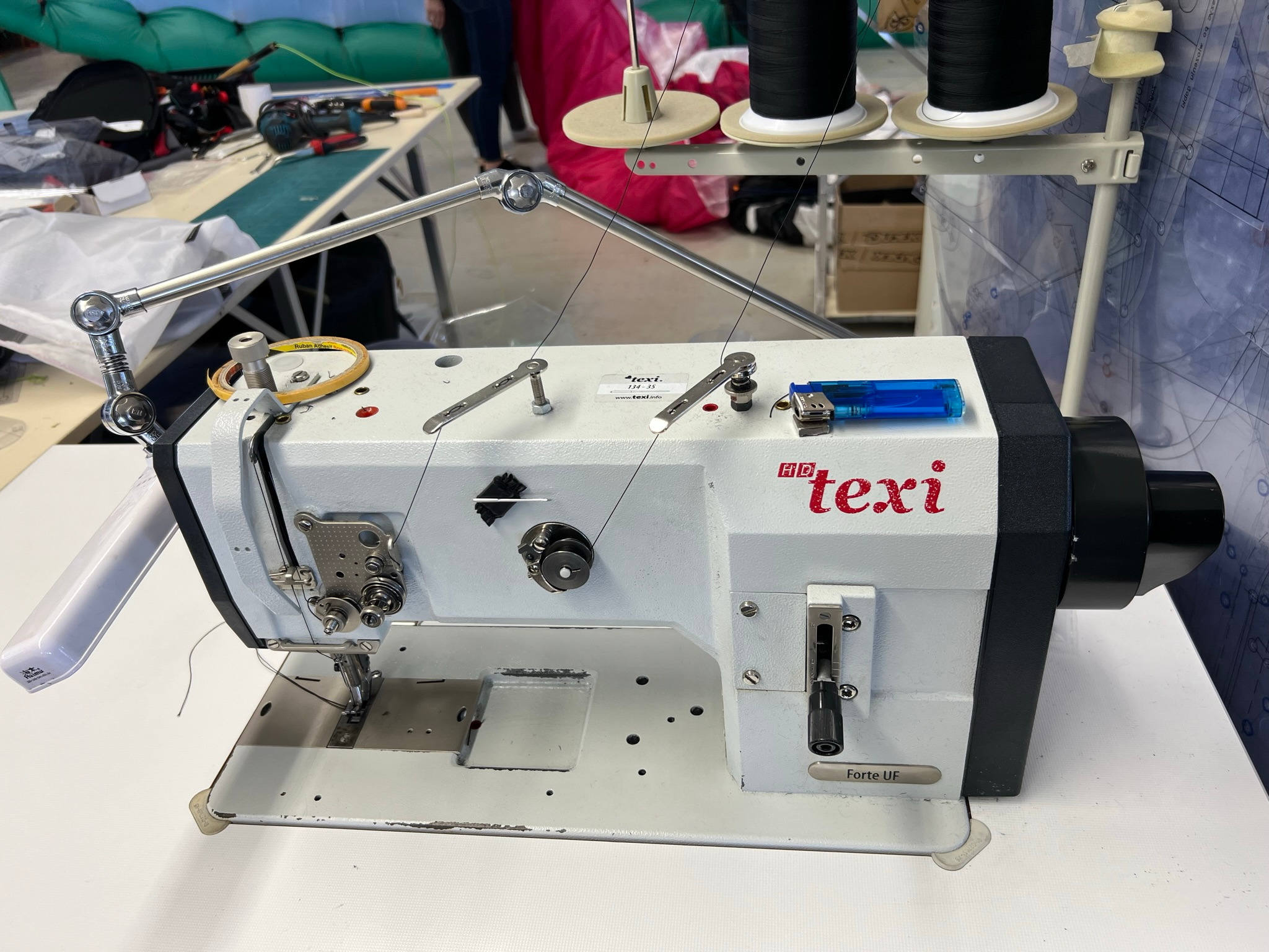 Profesjonalna maszyna do szycia Texi Forte UF z nawleczonymi czarnymi nićmi, oświetleniem i zapalniczką leżącą obok, na stole roboczym w szwalni.