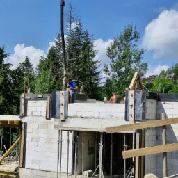 Wznoszenie ścian parteru z bloczków betonowych na fundamencie, widoczna pompa do betonu i pracownicy budowlani, w tle drzewa i zabudowa mieszkalna.