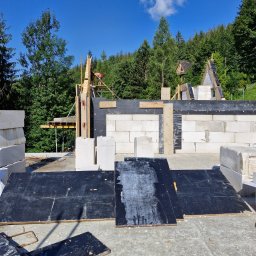 Usługi Minikoparką I Usługi Ogrodnicze Maks Aneta Kościelna - Wznoszenie ścian z bloczków betonu komórkowego na fundamencie w otoczeniu drzew, widoczny szalunek i pracownik na rusztowaniu.