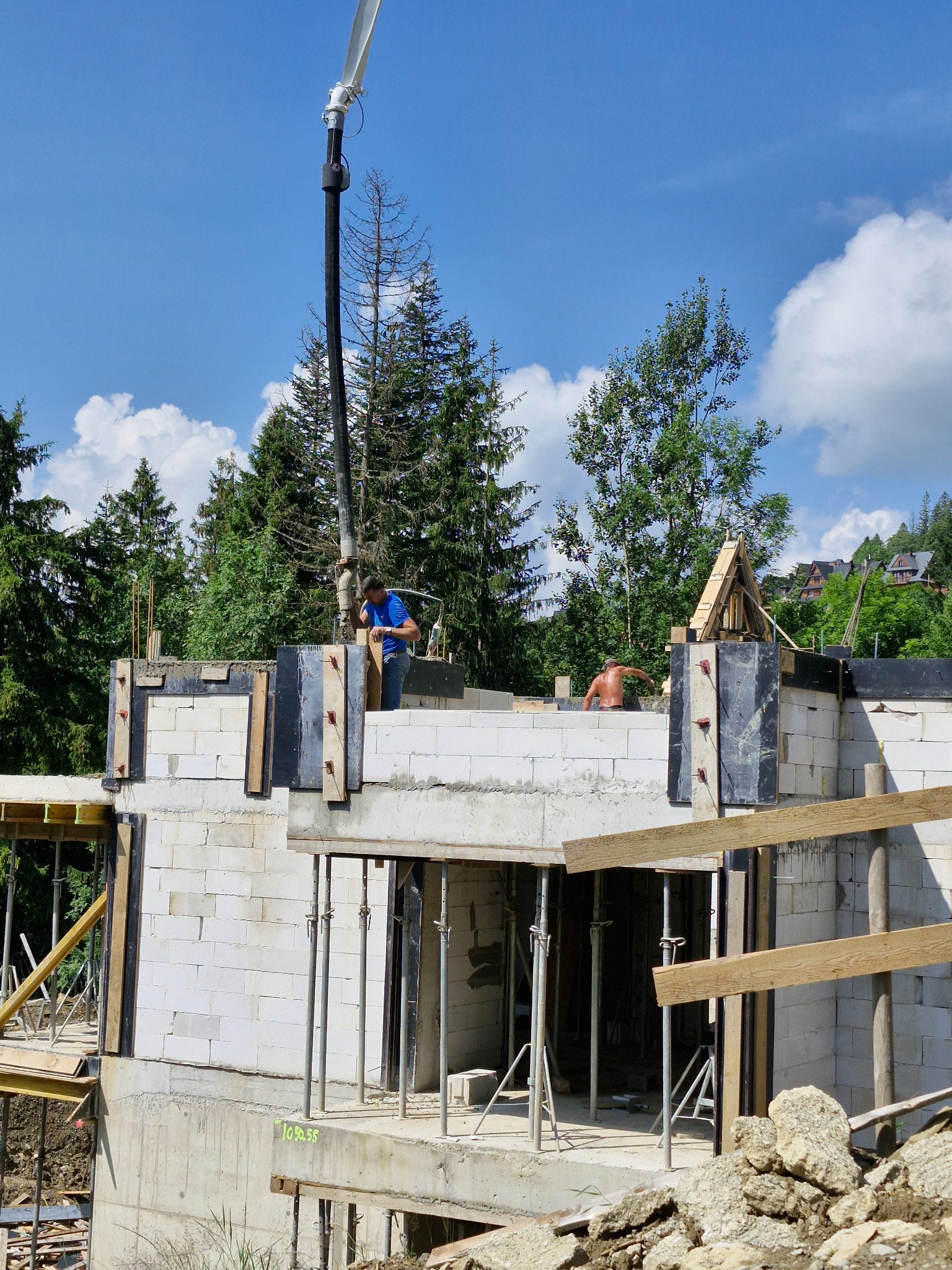 Wznoszenie ścian parteru z bloczków betonowych na fundamencie, widoczna pompa do betonu i pracownicy budowlani, w tle drzewa i zabudowa mieszkalna.