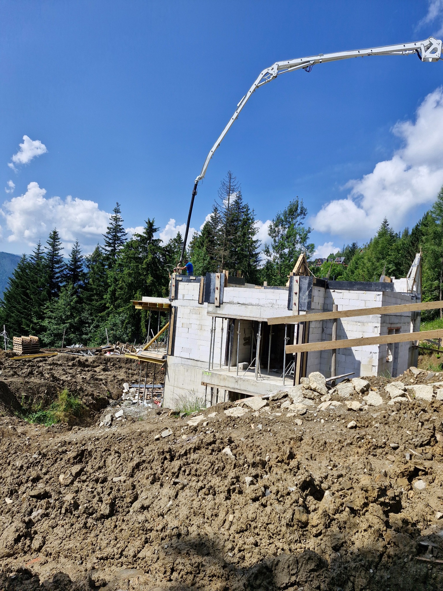 Budowa domu z betonu komórkowego na nierównym terenie, z widoczną pompą do betonu i stertą ziemi w wykopie.