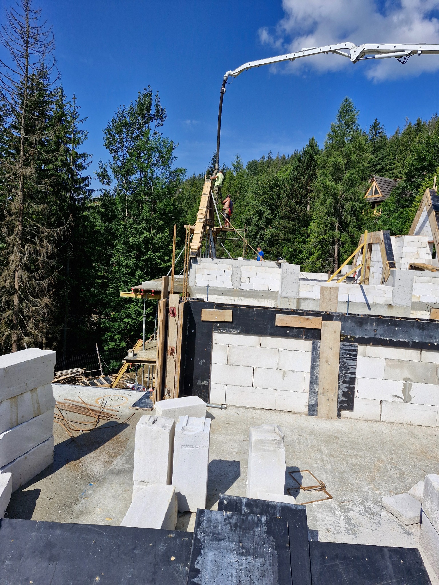 Wylewanie betonu za pomocą pompy do szalunków na budowie domu z białych bloczków, widoczne rusztowania, drewniane elementy konstrukcyjne i otaczający las.