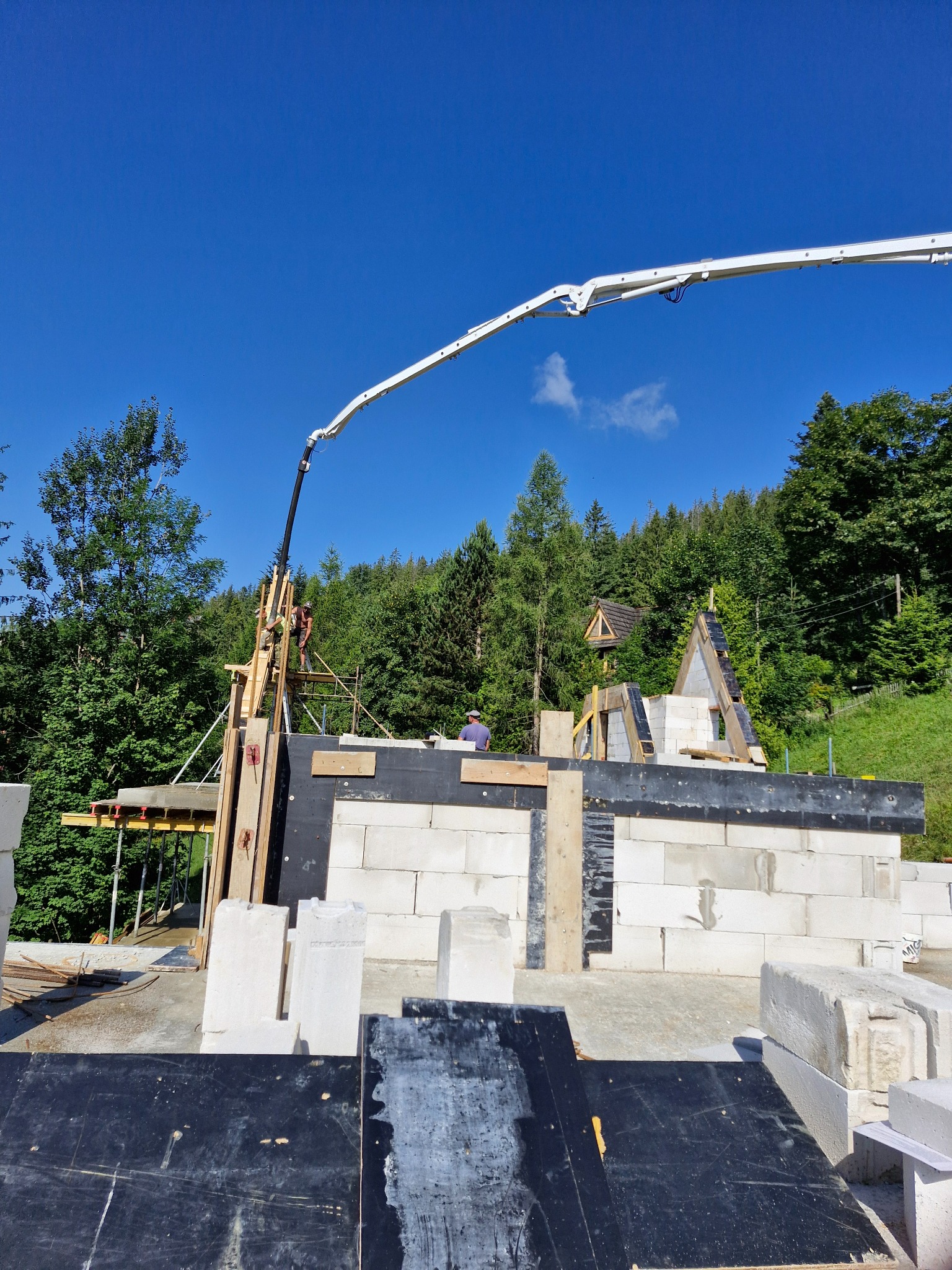 Wznoszenie ścian parteru domu z bloczków betonu komórkowego, widoczny wysięgnik pompy do betonu oraz pracownicy na rusztowaniu, w tle las iglasty i błękitne niebo.