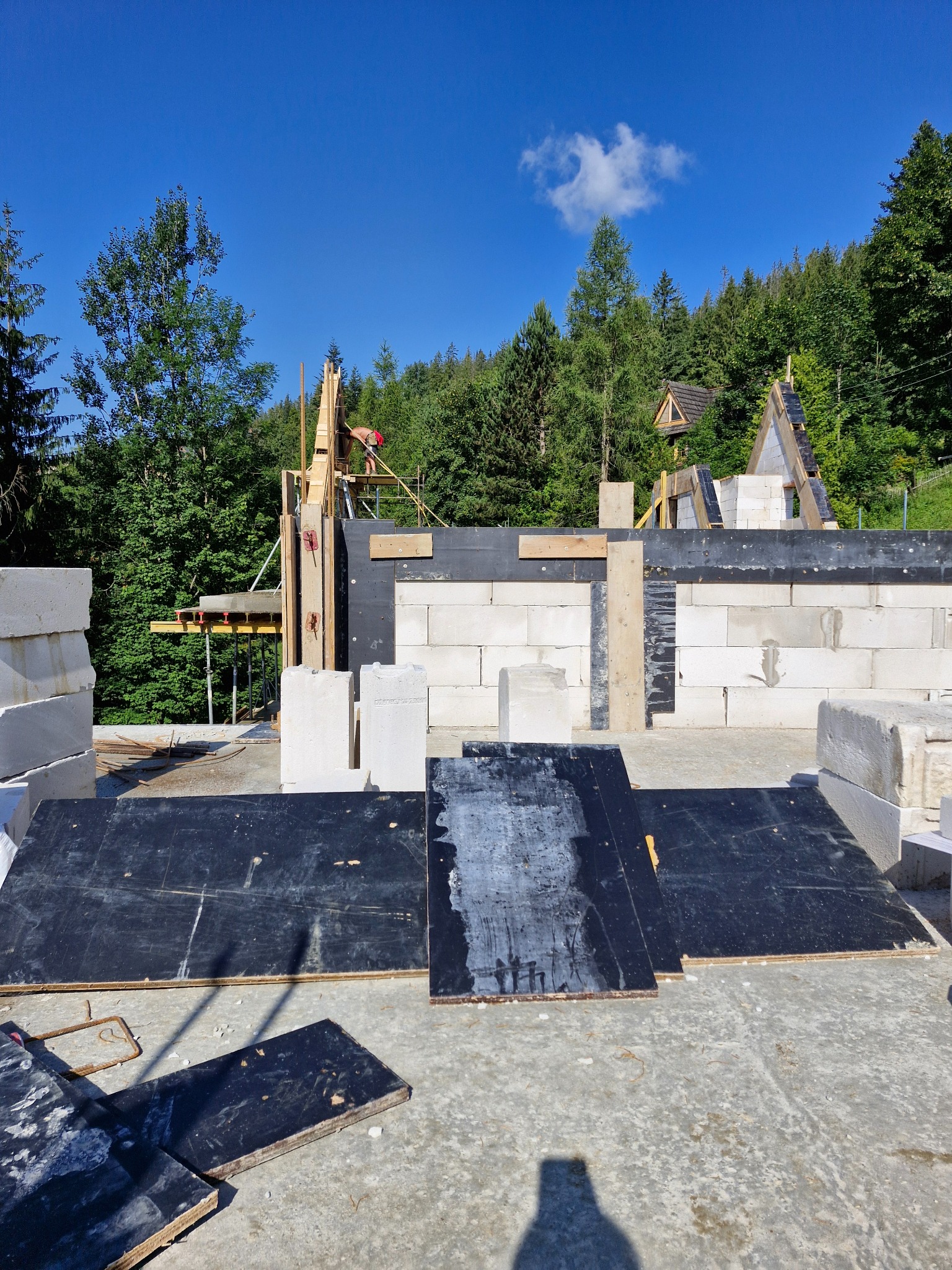 Wznoszenie ścian z bloczków betonu komórkowego na fundamencie w otoczeniu drzew, widoczny szalunek i pracownik na rusztowaniu.