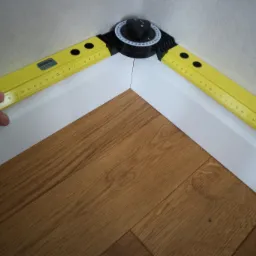 Pomiar kąta narożnika przypodłogowego za pomocą kątomierza i poziomicy, widoczna dłoń.