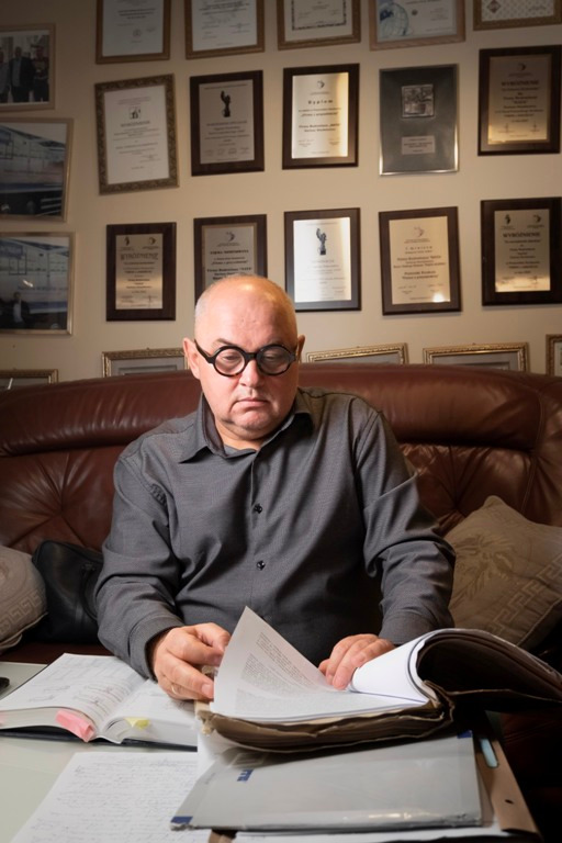 Mężczyzna w okularach siedzi na skórzanej kanapie, przeglądając dokumenty i otwarte księgi, na ścianie za nim wiszą liczne dyplomy i certyfikaty.