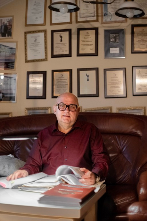 Portret mężczyzny w okularach siedzącego na skórzanym fotelu, przeglądającego otwartą książkę na białym stoliku, z certyfikatami i zdjęciami na ścianie w tle.