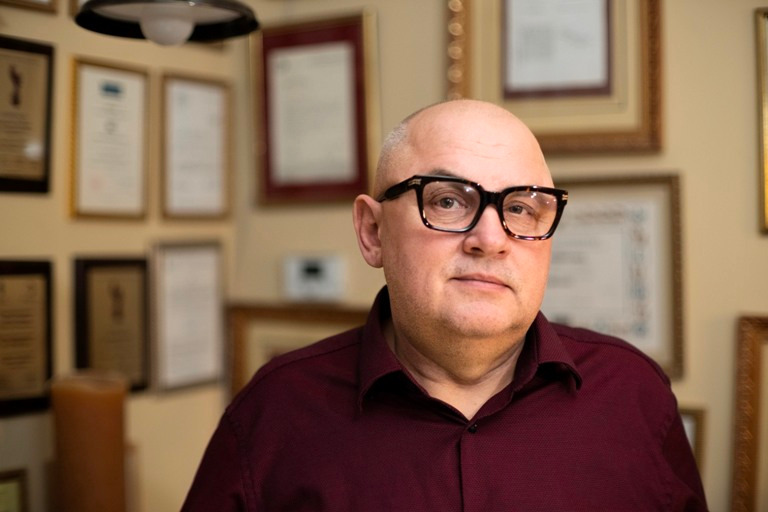 Portret mężczyzny w okularach na tle ściany z certyfikatami i nagrodami.