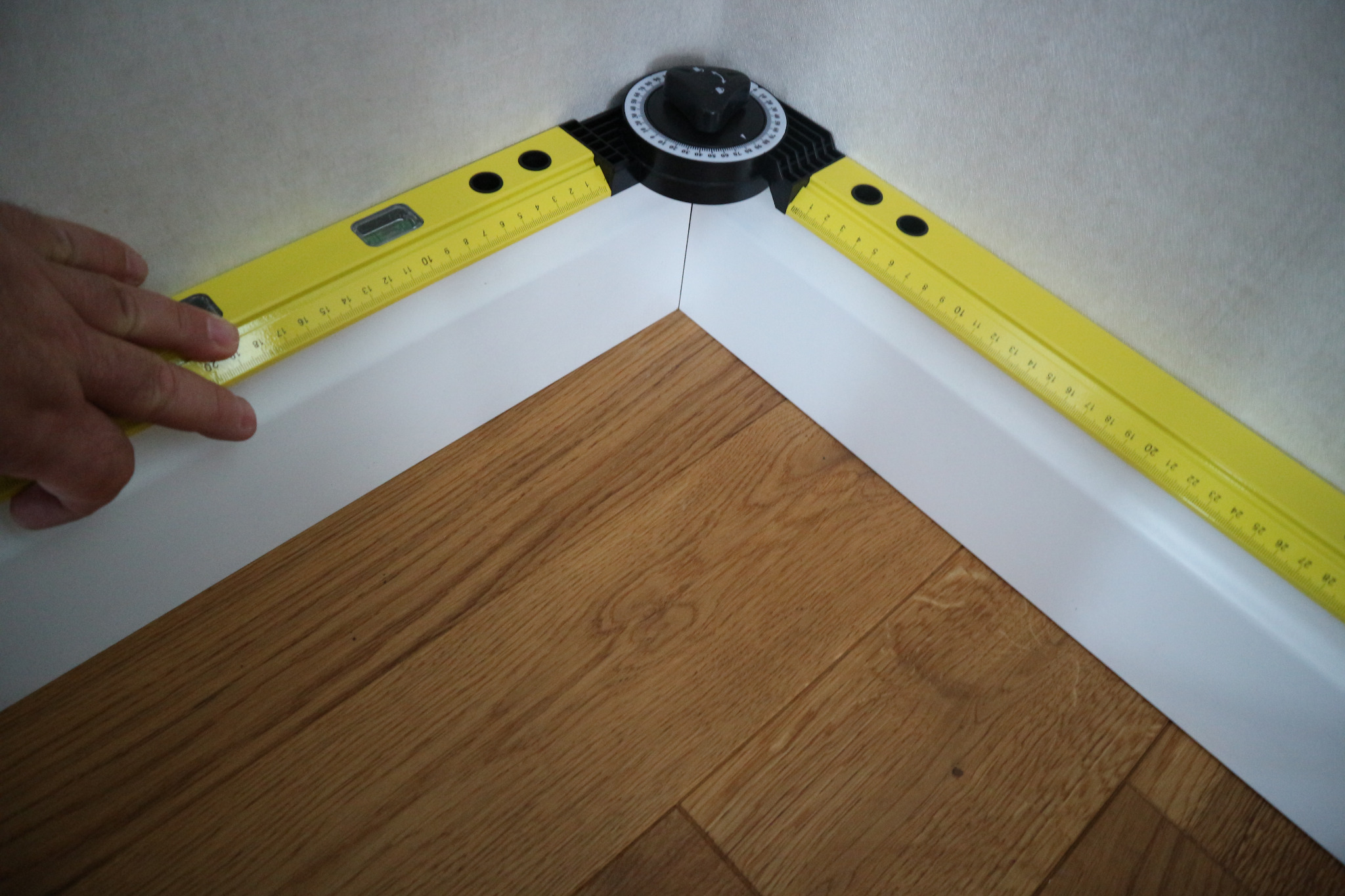 Pomiar kąta narożnika przypodłogowego za pomocą kątomierza i poziomicy, widoczna dłoń.